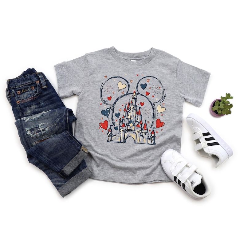 May include: A gray t-shirt featuring a castle design within a Mickey Mouse head outline, with red and blue hearts. The shirt is paired with blue jeans and white sneakers with black stripes. A small potted plant is also present.