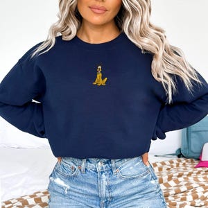 May include: Navy blue sweatshirt featuring a small embroidered Pluto design. The cartoon dog is depicted in yellow and black. The sweatshirt has a crew neck and long sleeves. The model is wearing blue jeans.