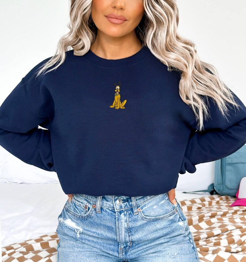May include: Navy blue sweatshirt featuring a small embroidered Pluto design. The cartoon dog is depicted in yellow and black. The sweatshirt has a crew neck and long sleeves. The model is wearing blue jeans.
