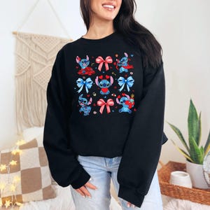 K&ouml;nnte beinhalten: Schwarzer Pullover mit einem blauen und roten Muster aus Comicfiguren, die Schleifen tragen. Die Figuren sind von Herzen und anderen dekorativen Elementen umgeben.