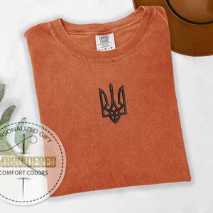 May include: A rust-colored t-shirt with a black embroidered Ukrainian trident symbol. The shirt is made of a soft, comfortable fabric and has a distressed look.