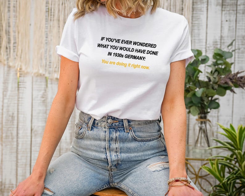 May include: White t-shirt with black text that reads "IF YOU'VE EVER WONDERED WHAT YOU WOULD HAVE DONE IN 1930s GERMANY: You are doing it right now."