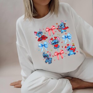 K&ouml;nnte beinhalten: Wei&szlig;er Pullover mit einer blauen Comicfigur, die ein rotes Herz tr&auml;gt und einen Bogen h&auml;lt. Die Figur wird im gesamten Design mit verschiedenen Posen und Accessoires wiederholt.