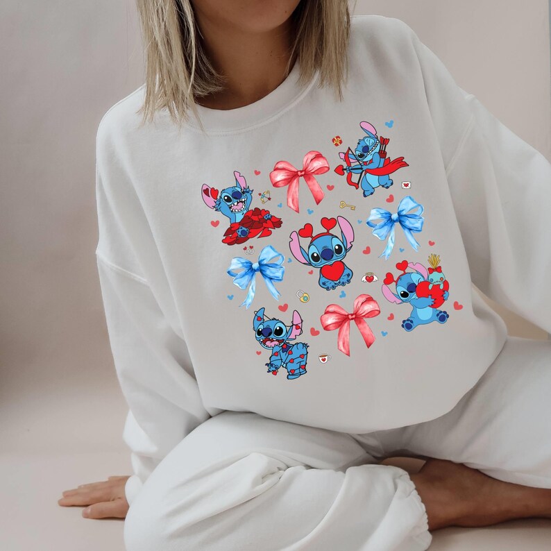 K&ouml;nnte beinhalten: Wei&szlig;er Pullover mit einer blauen Comicfigur, die ein rotes Herz tr&auml;gt und einen Bogen h&auml;lt. Die Figur wird im gesamten Design mit verschiedenen Posen und Accessoires wiederholt.