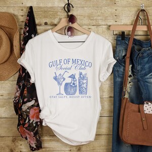 Puede incluir: Camiseta blanca con un gr&aacute;fico azul que dice "Gulf of Mexico Social Club" y presenta tres ilustraciones de c&oacute;cteles. El texto debajo del gr&aacute;fico dice "Stay Salty, Resist Often."
