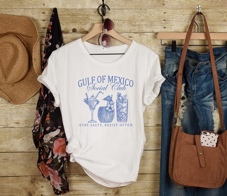 Puede incluir: Camiseta blanca con un gr&aacute;fico azul que dice "Gulf of Mexico Social Club" y presenta tres ilustraciones de c&oacute;cteles. El texto debajo del gr&aacute;fico dice "Stay Salty, Resist Often."