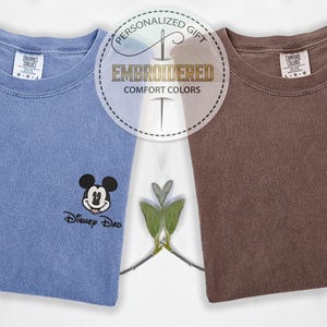 May include: Two folded t-shirts, one blue and one brown, each embroidered with a Disney character. The blue shirt features Mickey Mouse and the text "Disney Dad." The brown shirt has Minnie Mouse and "Disney Mom." The shirts are labeled "Embroidered Comfort Colors."