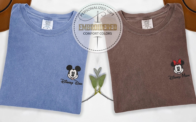 May include: Two folded t-shirts, one blue and one brown, each embroidered with a Disney character. The blue shirt features Mickey Mouse and the text "Disney Dad." The brown shirt has Minnie Mouse and "Disney Mom." The shirts are labeled "Embroidered Comfort Colors."