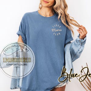 May include: A blue t-shirt with the text "Tired Moms Club" embroidered on the front. The shirt is being worn by a person with long blonde hair and a denim jacket.