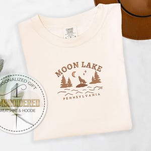 Pu&ograve; includere: Una maglietta bianca con un design grafico marrone che dice "Moon Lake Pennsylvania" con un lupo che ulula alla luna e alberi sullo sfondo.