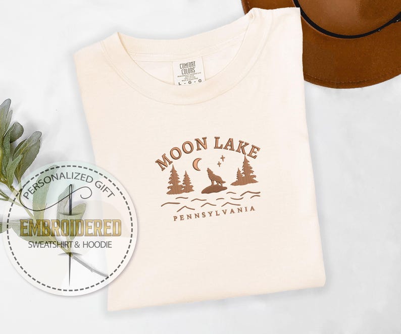 Pu&ograve; includere: Una maglietta bianca con un design grafico marrone che dice "Moon Lake Pennsylvania" con un lupo che ulula alla luna e alberi sullo sfondo.