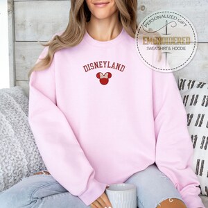 May include: Pink sweatshirt with the text "DISNEYLAND" in red and a red embroidered Mickey Mouse head with a bow.