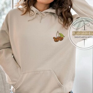 May include: Beige hoodie with a front pocket and drawstring hood. The left chest features an embroidered design of two jack-o'-lanterns on a cherry stem. A circular graphic reads "PERSONALIZED GIFT EMBROIDERED SWEATSHIRT & HOODIE."