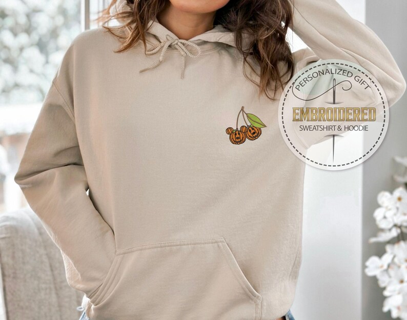 May include: Beige hoodie with a front pocket and drawstring hood. The left chest features an embroidered design of two jack-o'-lanterns on a cherry stem. A circular graphic reads "PERSONALIZED GIFT EMBROIDERED SWEATSHIRT & HOODIE."