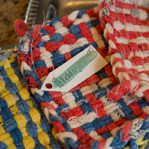 May include: Three woven pot holders in red, white, blue, and yellow. The pot holders are made of fabric strips and have a woven texture. One pot holder has a tag that says "Thread N' Yarn".