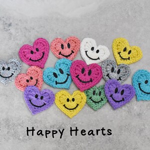 Peut inclure: Une collection de 12 ornements en forme de cœur en crochet avec des visages souriants. Les cœurs sont de différentes couleurs, notamment rose, bleu, jaune, vert et gris. Le texte "Happy Hearts" est écrit sous les cœurs.