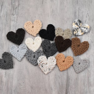 May include: A collection of crocheted hearts in various colors, including gray, brown, black, and beige. The hearts are arranged in a semi-circle on a gray wooden surface.