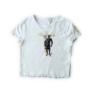May include: White crop top with a crew neck and ruffled edges. The shirt features a gold and black graphic of a figure with a staff and wings. The design is repeated on the sleeve. A casual, short-sleeved shirt.