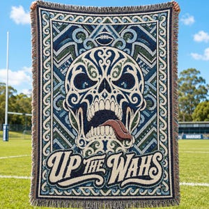 May include: A woven throw blanket featuring a large, intricate skull design with blue, green, and cream patterns. A football sits above the skull, and the text "UP THE WAHS" is printed in a bold, stylized font at the bottom. The blanket has fringed edges.