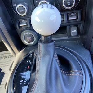 May include: A white leather-wrapped gear shift knob with black stitching and a black base. The knob has the numbers 2, 4, and R printed on it. The gear shift is surrounded by a black leather boot with brown stitching. The gear shift pattern is visible on the boot, with the letters P, R, N, and M-D printed on it.