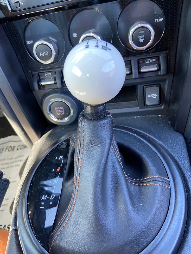 May include: A white leather-wrapped gear shift knob with black stitching and a black base. The knob has the numbers 2, 4, and R printed on it. The gear shift is surrounded by a black leather boot with brown stitching. The gear shift pattern is visible on the boot, with the letters P, R, N, and M-D printed on it.
