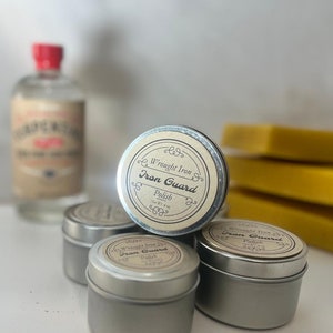 May include: Several small, silver metal tins of Wrought Iron Iron Guard Polish are stacked together. Each tin has a circular label with the product name. A bottle with a red cap and a label is in the background, along with yellow bars.