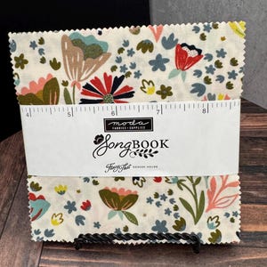 May include: A stack of square fabric pieces with a cream background and a multi-colored floral pattern. A white paper band wraps around the center, featuring the text Moda Fabrics Supplies Songbook Fancy That Design House and a ruler.