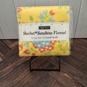 Può includere: Una pila di quadrati di tessuto in flanella gialla con un motivo floreale in blu, rosso e verde. Una fascia di carta bianca mostra il testo "moda FABRICS+SUPPLIES Sherbet*Sunshine Flannel by Corey Yoder of Coriander Quilts" e un righello.