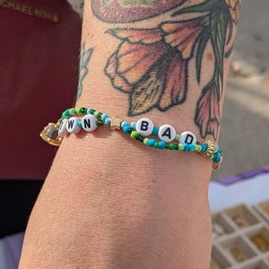 May include: A hand displays several rings, including a turquoise stone ring and an opal ring. A beaded bracelet spells out "OWN BAD" in white letter beads. The background shows jewelry displays.