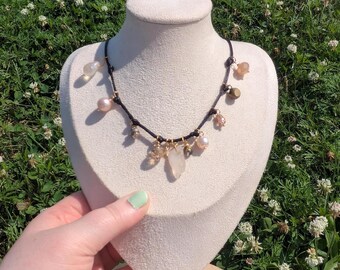 May include: A necklace with a black cord, featuring various beads and gemstones. The necklace includes pink, clear, and gold-colored beads, along with a large, clear quartz crystal pendant. The necklace is displayed on a cream-colored bust against a backdrop of green grass and white flowers.