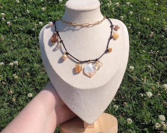 May include: A necklace display featuring two necklaces. The top necklace is a gold chain. The bottom necklace has a black cord with various beads, including pearls, amber-colored stones, and a white shell pendant. The display is on a cream-colored bust.