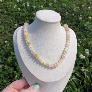 May include: A colorful beaded necklace displayed on a white bust. The necklace features a mix of pink, green, and yellow beads. The bust is on a wooden stand, set against a backdrop of green grass and white flowers.