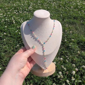 May include: A delicate necklace with a Y-shaped pendant, featuring small, colorful beads in pink, blue, and green. The necklace is displayed on a cream-colored bust against a backdrop of green grass and white flowers.