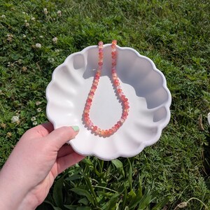 May include: A beaded necklace with a gradient of pink and orange gemstones rests in a white, scalloped dish. The dish is held by a hand with painted nails, set against a backdrop of green grass.