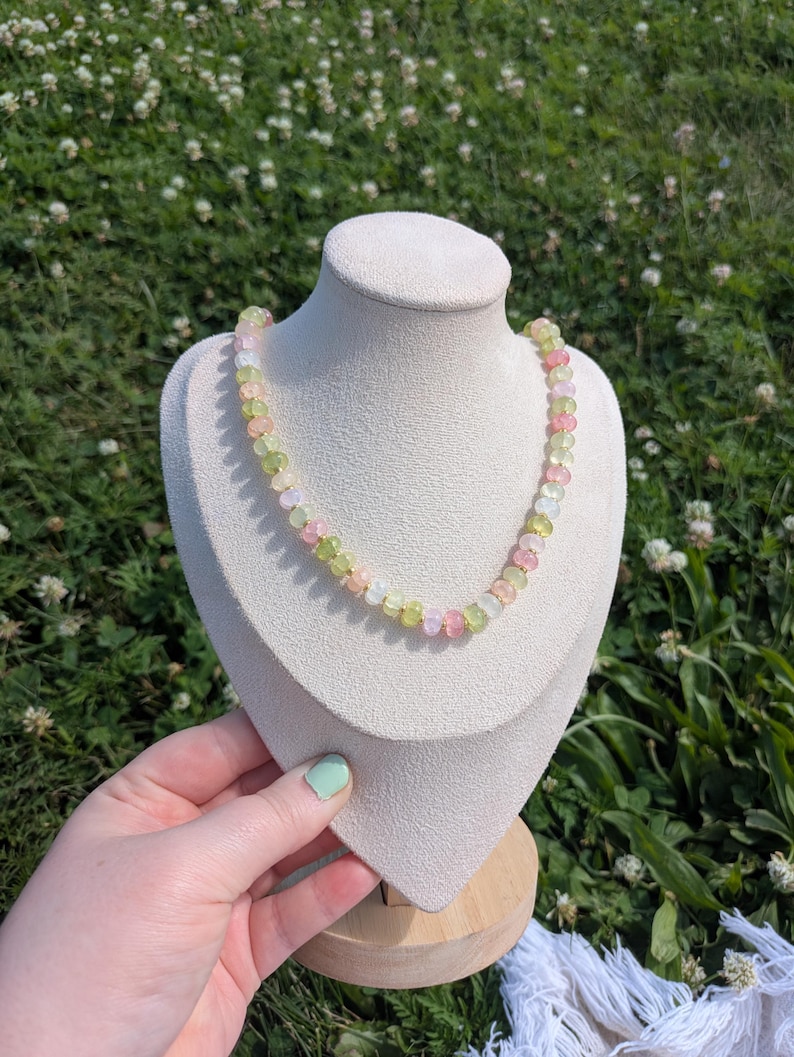 May include: A colorful beaded necklace displayed on a white bust. The necklace features a mix of pink, green, and yellow beads. The bust is on a wooden stand, set against a backdrop of green grass and white flowers.