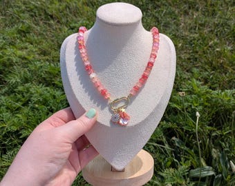 May include: A beaded necklace with pink and red gemstones. The necklace features a gold clasp and a charm with a flower and a pink teardrop-shaped stone. The necklace is displayed on a white jewelry bust.
