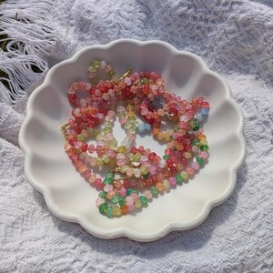 May include: A collection of colorful beaded necklaces in shades of pink, green, orange, and yellow, displayed in a white scalloped dish. The necklaces are made of small, round beads and are arranged on a white, textured surface.