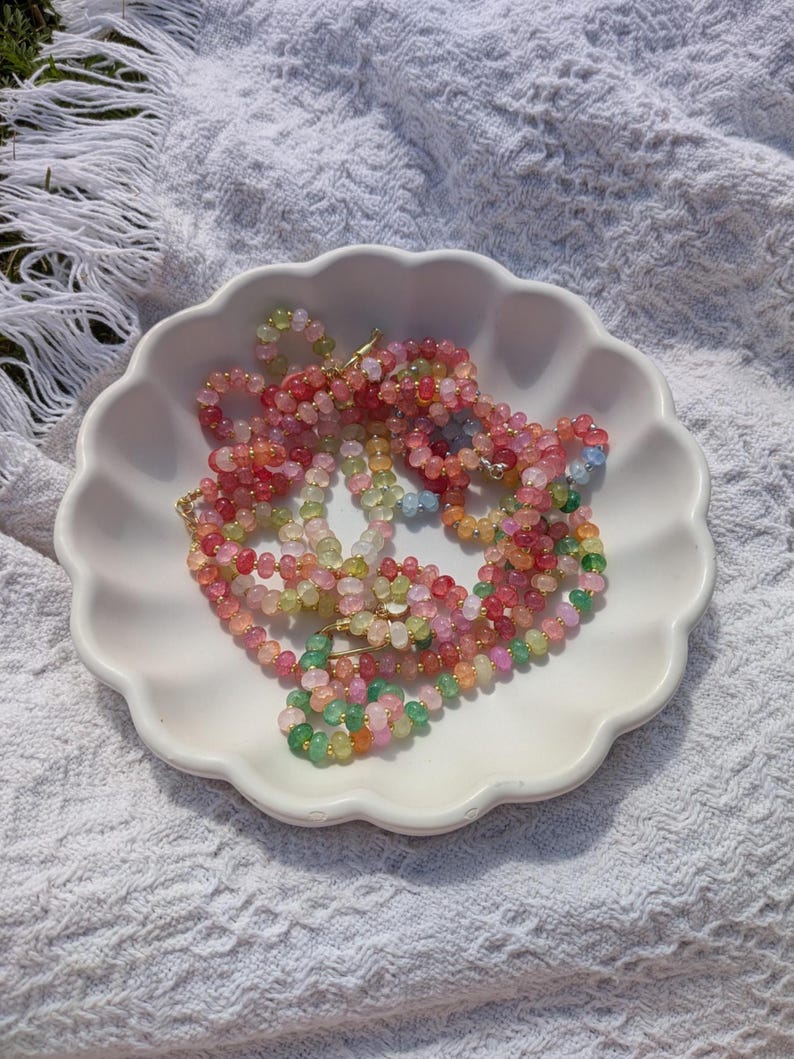 May include: A collection of colorful beaded necklaces in shades of pink, green, orange, and yellow, displayed in a white scalloped dish. The necklaces are made of small, round beads and are arranged on a white, textured surface.