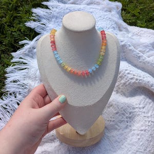 May include: A colorful beaded necklace displayed on a cream-colored bust. The necklace features a gradient of colors, including red, orange, yellow, green, and blue beads. The bust is set on a wooden base, with a white fringed cloth in the background.