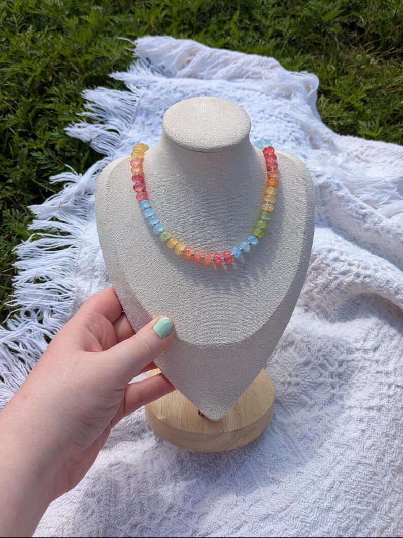 May include: A colorful beaded necklace displayed on a cream-colored bust. The necklace features a gradient of colors, including red, orange, yellow, green, and blue beads. The bust is set on a wooden base, with a white fringed cloth in the background.