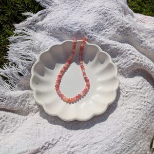 May include: A delicate necklace composed of small, round, pink and orange gemstones. The necklace rests on a white, scalloped-edge plate, set against a textured white blanket on a grassy background. The gemstones appear to be approximately 0.25 inches in diameter.