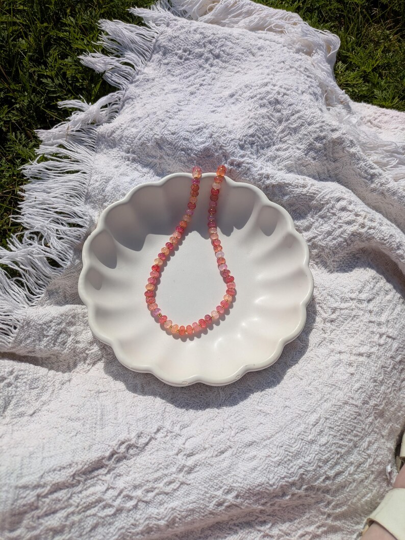 May include: A delicate necklace composed of small, round, pink and orange gemstones. The necklace rests on a white, scalloped-edge plate, set against a textured white blanket on a grassy background. The gemstones appear to be approximately 0.25 inches in diameter.