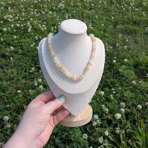May include: A multi-colored gemstone necklace displayed on a white bust. The necklace features a variety of pastel-colored beads, including pink, green, and yellow. The bust is set against a backdrop of green grass and white flowers.