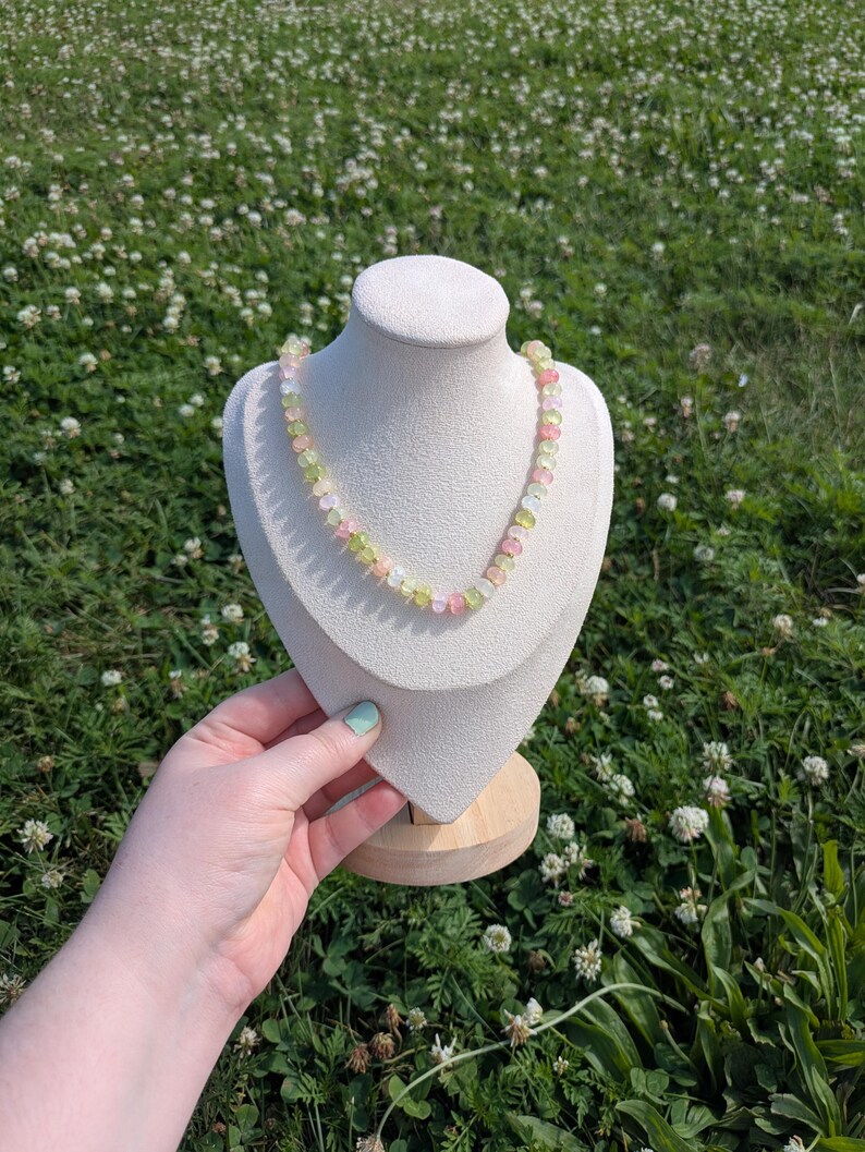 May include: A multi-colored gemstone necklace displayed on a white bust. The necklace features a variety of pastel-colored beads, including pink, green, and yellow. The bust is set against a backdrop of green grass and white flowers.