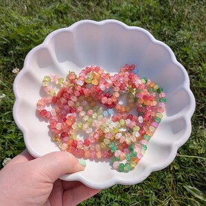 May include: A white, scalloped-edge bowl filled with colorful beaded necklaces. The necklaces feature a variety of colors, including pink, red, green, yellow, and blue. The beads are small and round, and the necklaces appear to be handmade. The bowl is held by a person with a green painted fingernail.