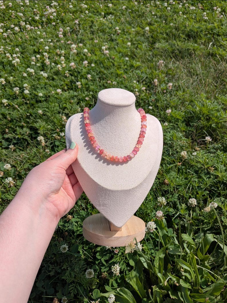 May include: A pink and orange beaded necklace displayed on a white bust against a backdrop of green grass and white flowers. The necklace is composed of small, round beads in varying shades of pink and orange.