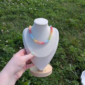May include: A colorful beaded necklace displayed on a white bust against a backdrop of green grass. The necklace features a rainbow of colors, including red, orange, yellow, green, and blue beads. The bust is on a wooden stand.