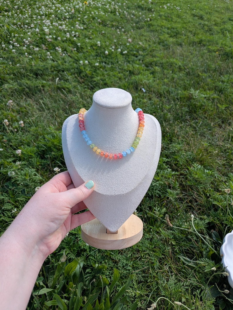 May include: A colorful beaded necklace displayed on a white bust against a backdrop of green grass. The necklace features a rainbow of colors, including red, orange, yellow, green, and blue beads. The bust is on a wooden stand.