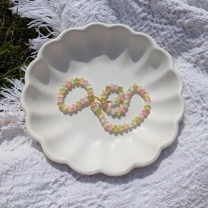 May include: A delicate necklace featuring a strand of small, round beads in shades of pink, green, and yellow. The necklace is displayed on a white, scalloped-edge dish, set against a textured white background. The necklace has a gold-colored clasp.