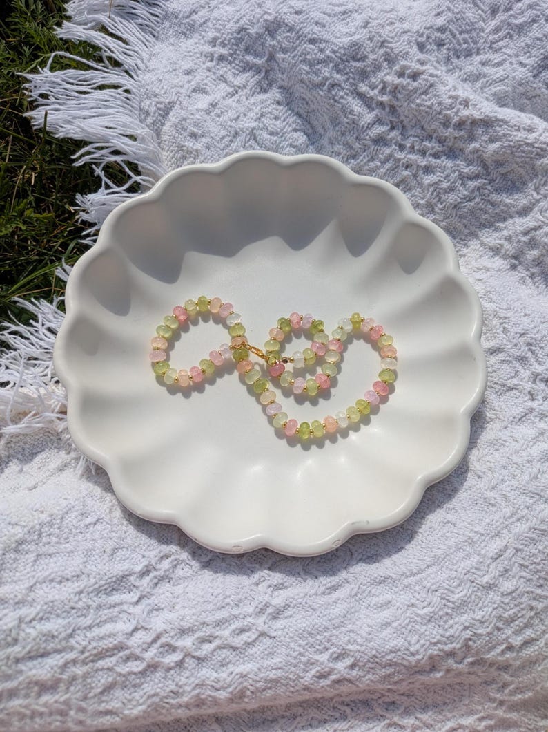 May include: A delicate necklace featuring a strand of small, round beads in shades of pink, green, and yellow. The necklace is displayed on a white, scalloped-edge dish, set against a textured white background. The necklace has a gold-colored clasp.